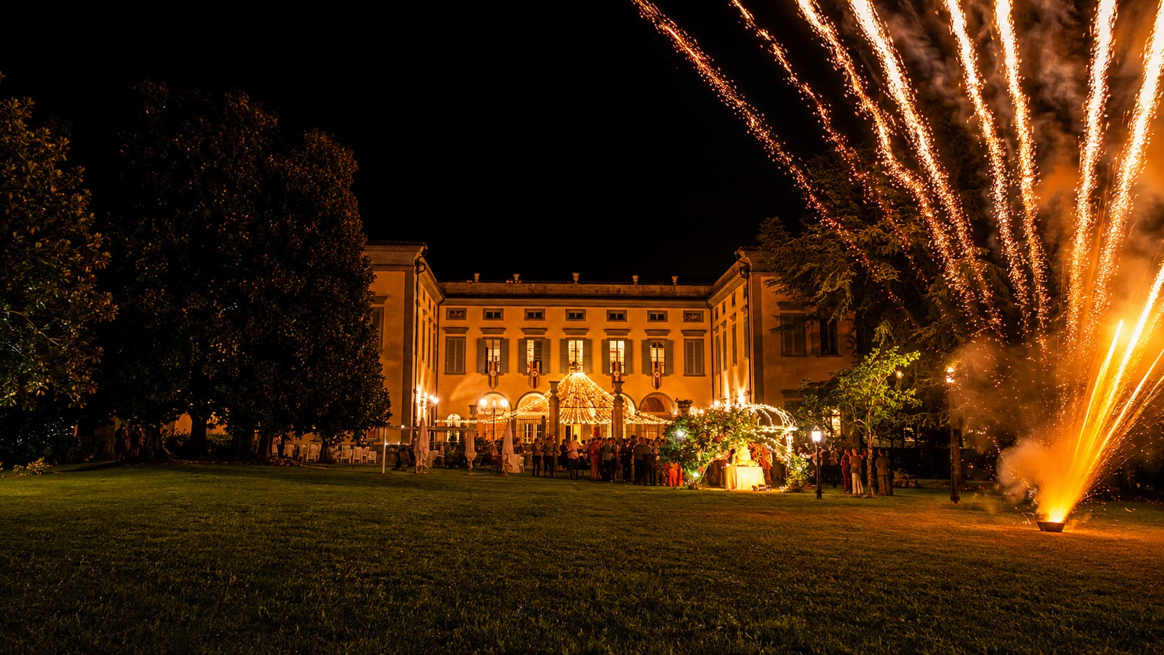 La facciata della villa illuminata di notte, con i fuochi d'artificio che salgono sopra il tetto